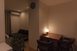 Una habitación pequeña y con poca luz, con una cama, un escritorio con dos sillas, un reloj en la pared y una planta en maceta.