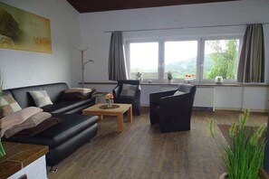 Ein Wohnzimmer mit einer Couch, einem Couchtisch und einem Sessel. Es gibt Fenster mit Vorhängen, eine Pflanze und ein Bild an der Wand.
