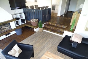 A living room with a TV, seating, a staircase, and a kitchen in the background.
