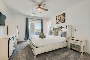 Een slaapkamer met een bed, een nachtkastje, een plafondventilator, een raam met gordijnen en een aan de muur bevestigde televisie.