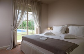 Ein Schlafzimmer mit einem großen Bett, einer Nachttischlampe und Blick auf einen Swimmingpool durch ein Fenster.