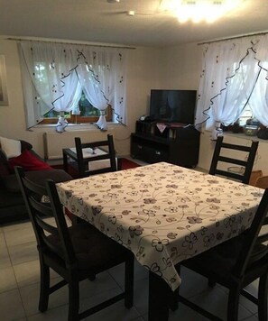 Ein Wohnzimmer mit Esstisch, Stühlen, einer Couch, einem Fernseher und Fenstern mit Vorhängen.