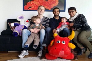 Eine Familie sitzt auf einem Sofa, im Hintergrund hängt ein Bild und ein großes Stofftier ist zu sehen.