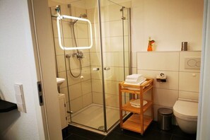 A modern bathroom with a glass-enclosed shower, a wooden shelf with towels, a toilet, and a small trash bin. - New Koralle Port in Bremerhaven (Bremerhaven)