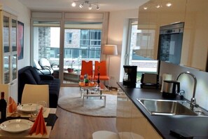 A modern living room with a sofa, dining table, and chairs, a kitchen with a sink and appliances, and a view through large windows. - New Koralle Port in Bremerhaven (Bremerhaven)