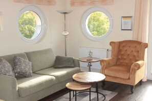 A room with a sofa, armchair, round tables, and two windows with a view of greenery.