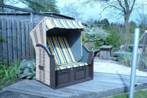 Ein Korbstuhl mit gestreifter Sitzkissen, ein Holzdeck und ein Metallzaun in einer Gartenanlage.