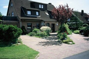 Ein zweistöckiges Haus mit Backsteinfassade, geneigtem Dach und Hauseingang. Ein gepflasterter Weg führt zum Eingang, und das Anwesen ist von gepflegten Gärten und Sträuchern umgeben.