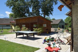 Uma cabana de madeira com mesa e cadeiras, cercada por vegetação e uma área de recreação com brinquedos.