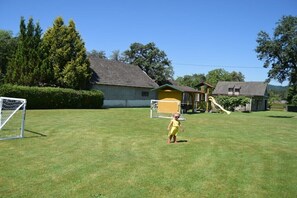Um gramado com um gol de futebol, um pequeno edifício amarelo e uma casa maior com telhado inclinado.