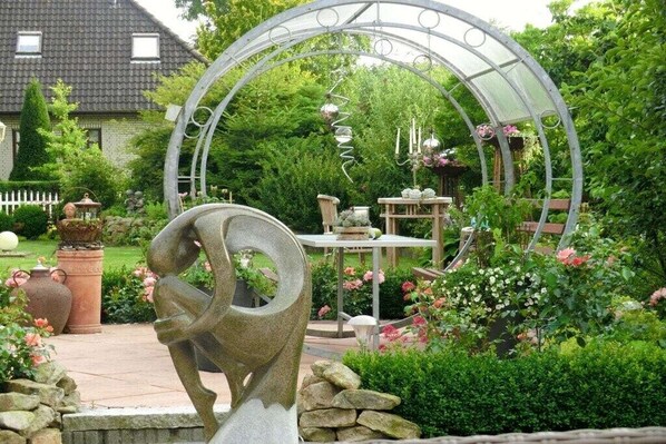 Ein Garten mit einer Steinskulptur, einem Metallbogen und einem Tisch mit Blumen.