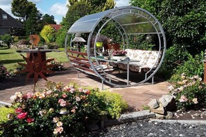 Ein Garten mit Gewächshaus, einem Holztisch mit Dekoration und verschiedenen Pflanzen und Blumen.