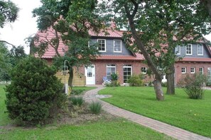 Ein zweistöckiges Backsteinhhaus mit rotem Dach, einer Haustür und einem kleinen Vorgarten mit Weg und Baum.