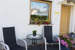 Ein kleiner Sitzbereich im Freien mit zwei Stühlen und einem Tisch, ein Fenster mit Blumen und eine Tür.