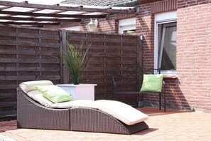 A wicker lounge chair with cushions, a potted plant, and a brick wall with a window. - Holiday home in Hage with swimming pool (Hage)