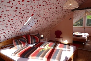 Ein Schlafzimmer mit schrägen Wänden, einem Bett mit gestreifter Bettwäsche, einem Nachttisch mit Lampe und einem Fenster mit Blick ins Grüne.