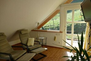 Ein Zimmer mit schrägen Wänden, zwei Stühle, ein kleiner Tisch und ein Fenster mit Blick auf eine grüne Landschaft.
