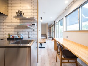 Eine moderne Küche mit Spüle aus Edelstahl, einem Wasserkocher und einer Kaffeemaschine. Ein Holztisch mit Stühlen lädt zum Verweilen ein. Die Küche verfügt über helle Ziegelwände und ein Fenster, das für natürliches Licht sorgt.