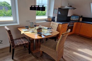 Un coin repas avec une table dressée pour quatre personnes, des chaises en osier, une cuisine équipée d’une cuisinière et d’un four, et une fenêtre donnant sur un paysage verdoyant.