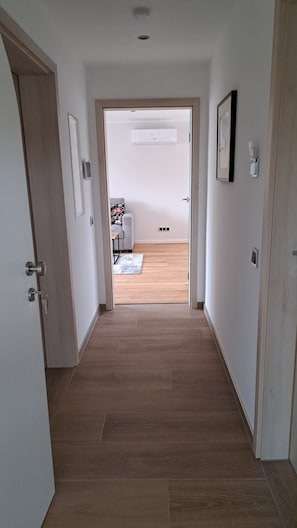 Een hal met houten vloer, een deur aan de linkerkant en een kamer met een bank en vloerkleed op de achtergrond.