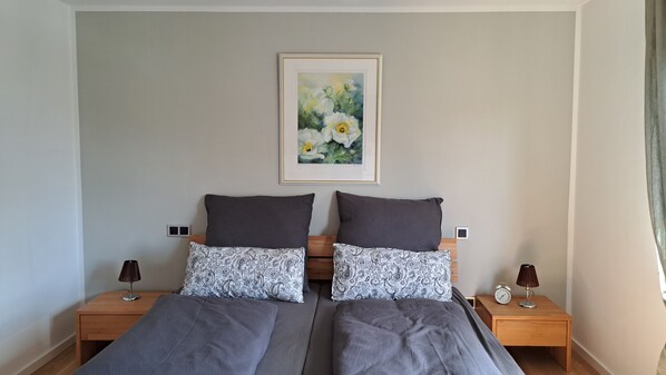 Un lit double avec une literie grise, deux tables de chevet avec des lampes, un tableau floral encadré accroché au mur et une tête de lit en bois.
