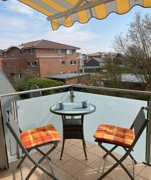 Ein Balkon mit einem Glastisch und zwei Stühlen, Blick auf Gebäude im Hintergrund und ein gelbes Markisenvorzelt.