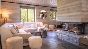 Ein modernes Wohnzimmer mit Ecksofa, rundem Couchtisch, Hocker, Kamin und großen Fenstern.