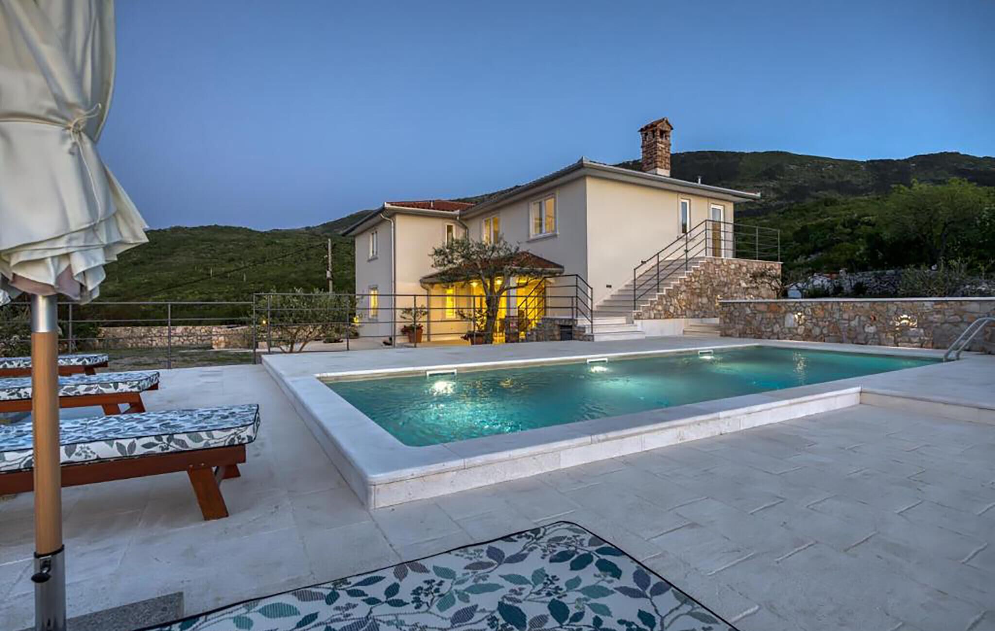 Un espace aménagé au bord de la piscine, avec des chaises longues, une piscine et une maison dotée d’un escalier en pierre et d’une terrasse.