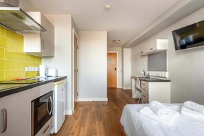 A compact hotel room with a kitchenette, a bed with white linens, a TV, and a wooden floor.