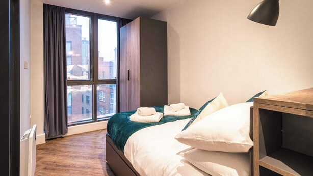 Ein Schlafzimmer mit großem Fenster, einem Bett mit weißer Bettwäsche, einem Nachttisch aus Holz und einer modernen Lampe.