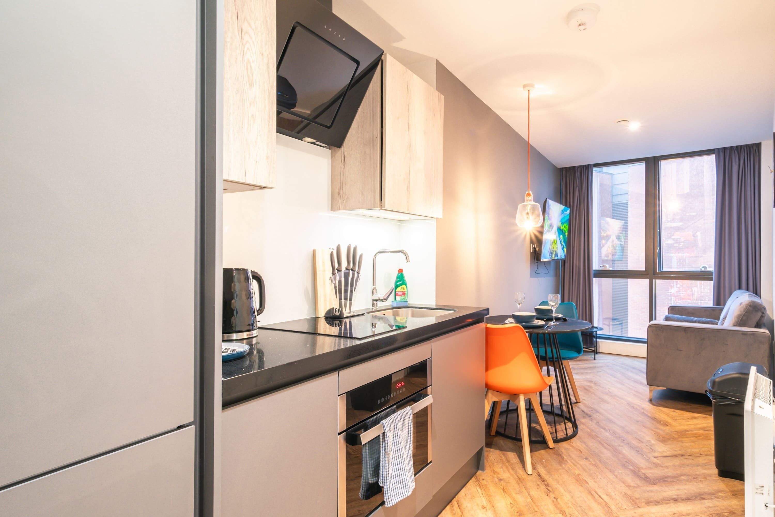 A modern kitchen with a built-in oven, a dining area with a table and chairs, and a sofa in a living area with a large window.