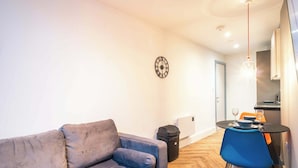 Ein modernes Wohnzimmer mit einem grauen Sofa, einem blauen Sessel, einem Esstisch mit schwarzem Tischläufer und einer Wanduhr.