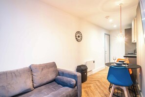 Ein modernes Wohnzimmer mit einem grauen Sofa, einem blauen Sessel, einem Esstisch mit schwarzem Tischläufer und einer Wanduhr.