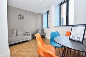 Ein modernes Zimmer mit grauem Sofa, orangefarbenen und blauen Sesseln, einem runden Tisch, einer Wanduhr und einem Fenster mit Jalousien.