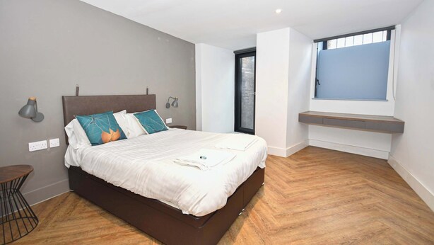 Un dormitorio moderno con una cama grande, una mesita redonda pequeña, un banco empotrado y un ventanal con persianas.
