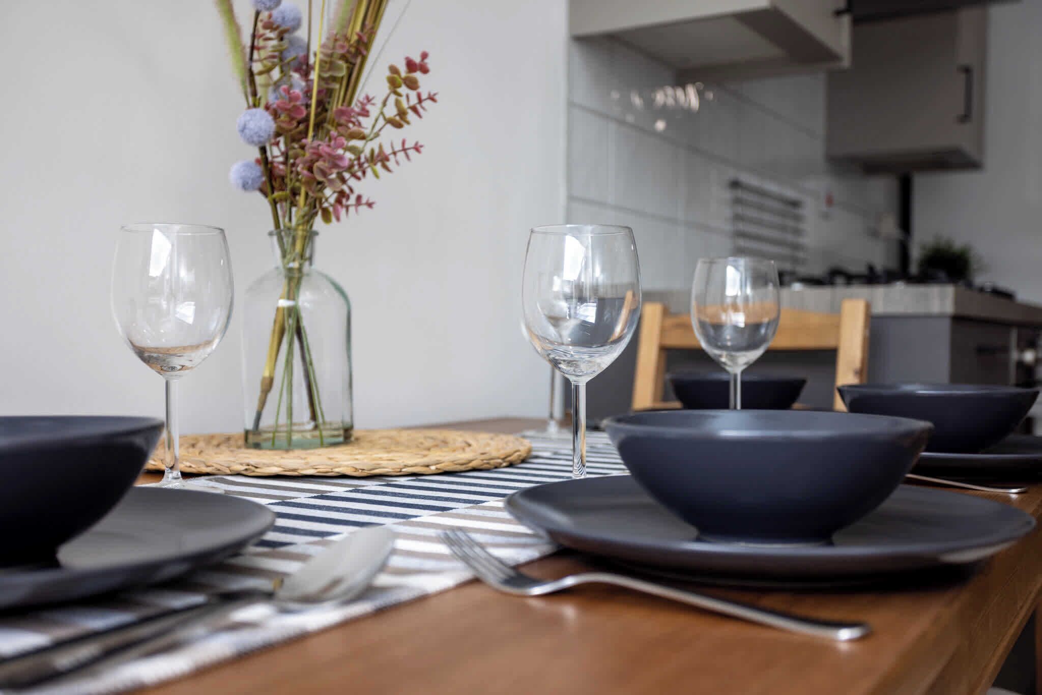 A dining table set with plates, bowls, cutlery, and wine glasses, with a vase of flowers in the background.