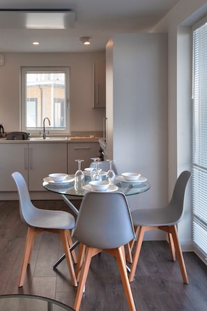 Eine moderne Küche mit Essbereich, inklusive Glastisch, grauen Stühlen und einem Fenster mit Jalousien.