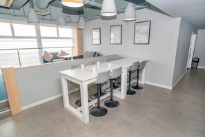 A modern kitchen with a white countertop, bar stools, pendant lights, and a view through large windows. - Central Coventry Budget Studio w Gym & Games Room (Coventry)