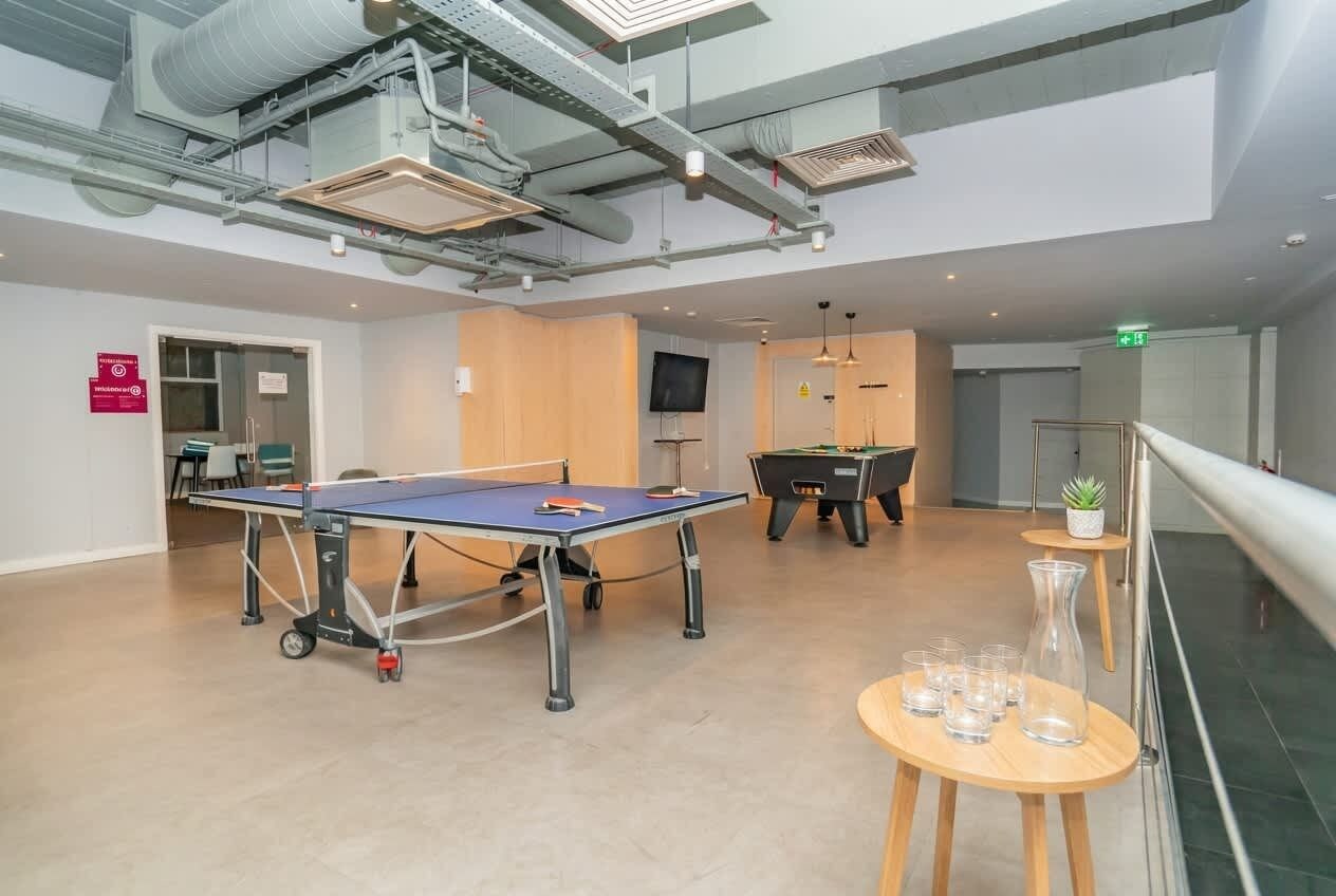 A spacious indoor area with a ping pong table, a pool table, and a billiards table. There are chairs and a small table with glasses.