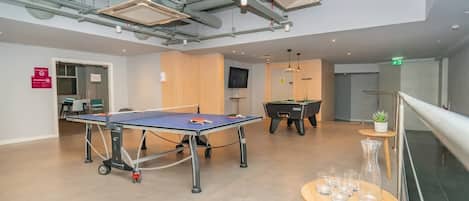 A spacious indoor area with a ping pong table, a pool table, and a billiards table. There are chairs and a small table with glasses.