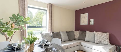 A modern living room with a dining area, a grey sofa, and a large window with curtains.
