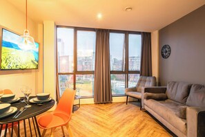 Ein modernes Wohnzimmer mit einer Couch, einem Esstisch mit Stühlen, einem an der Wand montierten Fernseher und großen Fenstern mit Vorhängen.
