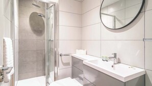 Een moderne badkamer met een douche met glazen wand, een wit wastafelmeubel met toiletartikelen, en een ronde spiegel.