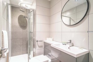 Ein modernes Badezimmer mit einer gläsernen Duschkabine, einem weißen Waschbecken mit Pflegeprodukten und einem runden Spiegel an der Wand.