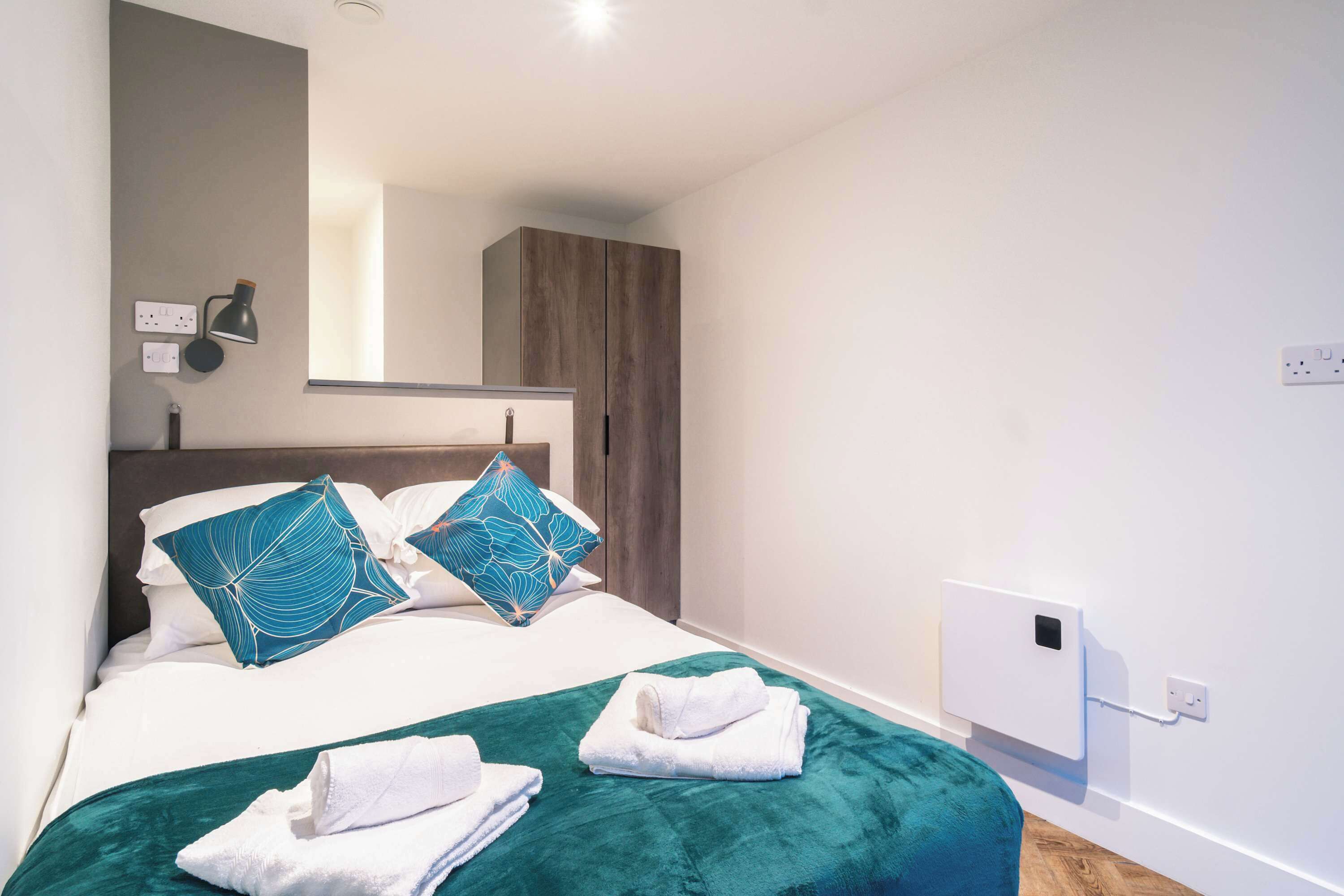 Habitación de hotel con cama, dos almohadas, una manta verde, dos toallas dobladas, una mesita de noche, una lámpara, un espejo, un armario y un enchufe.