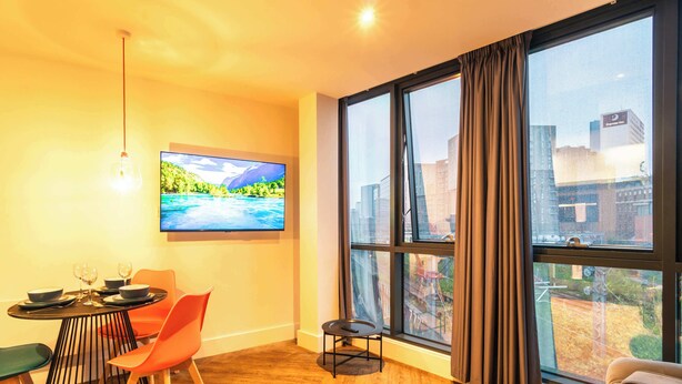 Een kamer met een eettafel, stoelen, een televisie en een groot raam met gordijnen.