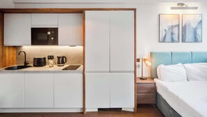 A modern kitchen with a microwave, toaster, and coffee maker, a sink, and a bed with a blue headboard. - Modern Studio Apartment in Central Hastings (East Sussex)