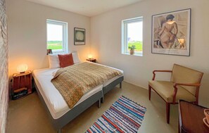 Ein Schlafzimmer mit einem Bett, Nachttischlampen, einem Sessel, einem Teppich und zwei Fenstern mit Kunstwerken.