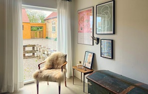 Ein Zimmer mit Blick durch ein Fenster, ein Holzstuhl, ein kleiner Tisch mit einer Glasflasche und gerahmte Kunstwerke an der Wand.
