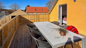 Eine Terrasse mit Tisch und Stühlen, ein Holzzaun und im Hintergrund ein Gebäude mit rotem Dach.